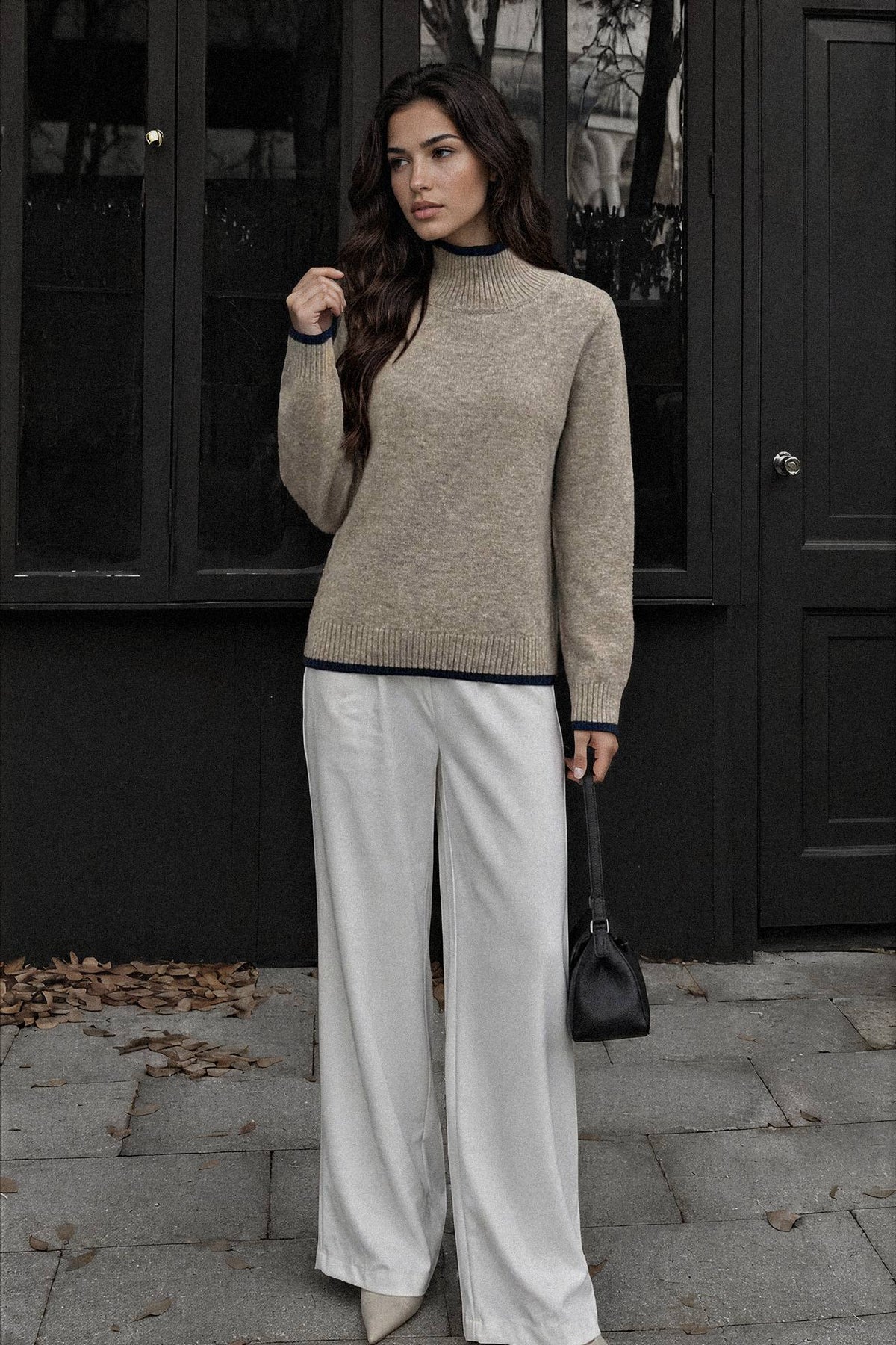A woman stands outdoors in front of a dark building, wearing a beige knit mock-neck sweater with dark trim, white wide-leg pants, and holding a small black handbag.