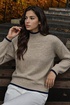 A woman wearing a beige knit mock-neck sweater with dark navy ribbed trim at the collar, cuffs, and hem, paired with white pants, sitting on outdoor wooden steps with autumn foliage in the background.