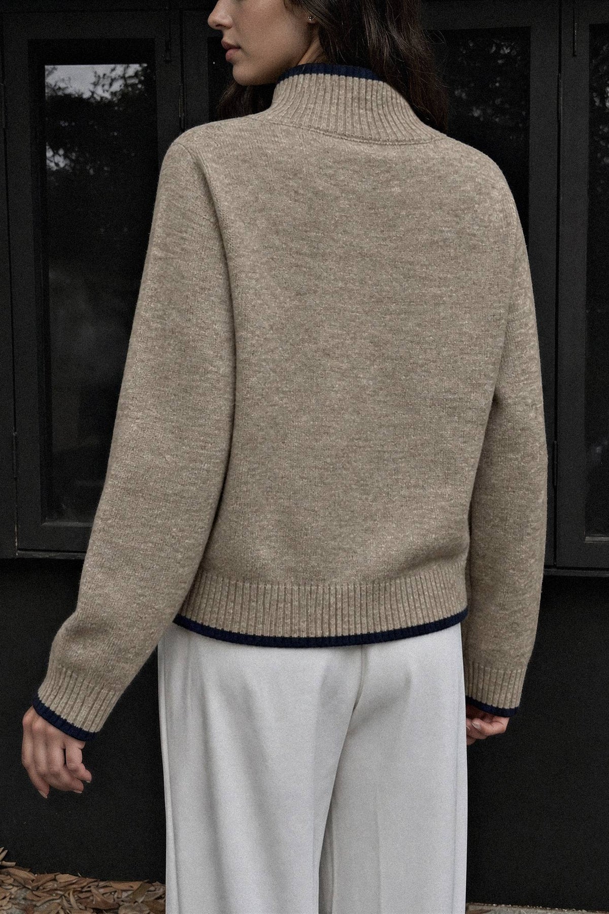 A woman shown from the back wearing a beige knit mock-neck sweater with dark navy ribbed trim at the collar, cuffs, and hem, paired with white trousers, set against a dark windowed background.