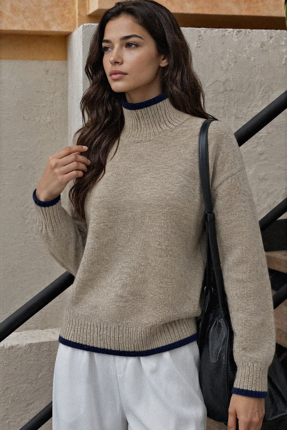 A woman wearing a beige knit mock neck sweater with navy blue ribbed trim, paired with white trousers and a black shoulder bag, standing on outdoor stone steps.