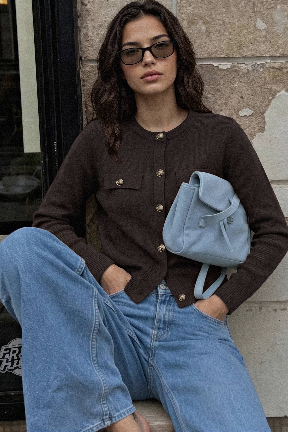 A woman wearing a dark brown button-up cardigan, light blue jeans, and a light blue backpack sits against a stone wall.