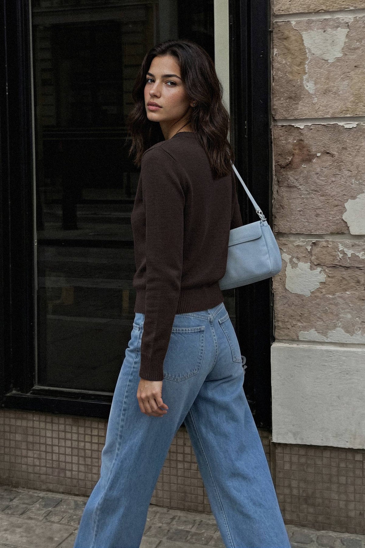A woman wearing a dark brown long-sleeve knit sweater and wide-leg light blue jeans, carrying a light blue shoulder bag, walking outdoors next to a stone wall.