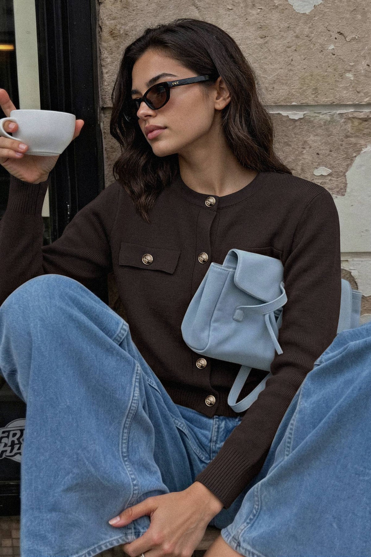 A woman wearing a dark brown button-up cardigan, light blue wide-leg jeans, and a small light blue shoulder bag sits against a stone wall, holding a white coffee cup.