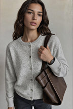 A woman wearing a light gray button-up cardigan sweater and dark jeans, carrying a brown leather shoulder bag against a neutral beige background.