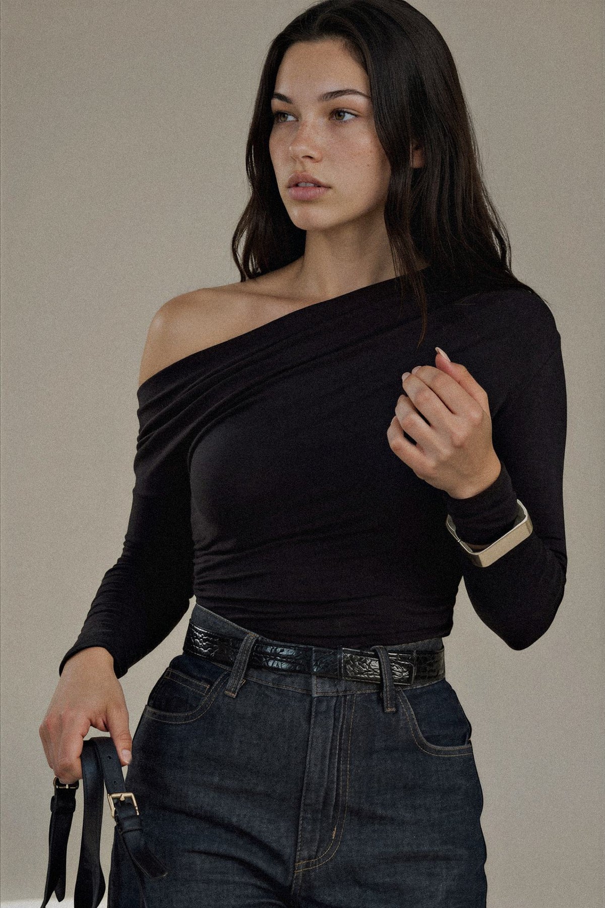A woman modeling a black long-sleeve off-the-shoulder top paired with dark blue jeans and a black belt, holding a black strap against a plain beige studio background.