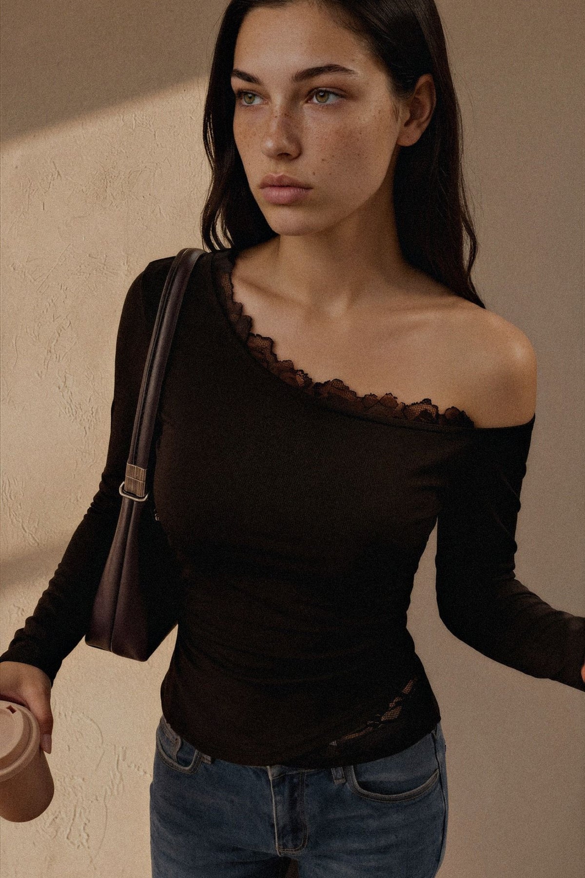 A woman wearing a dark brown off-the-shoulder long-sleeve top with lace trim, paired with blue jeans and carrying a brown shoulder bag, set against a textured beige wall.