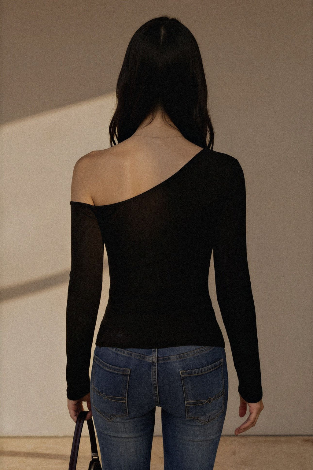A woman shown from behind wearing a black long-sleeve one-shoulder top and blue jeans, standing against a plain beige wall.