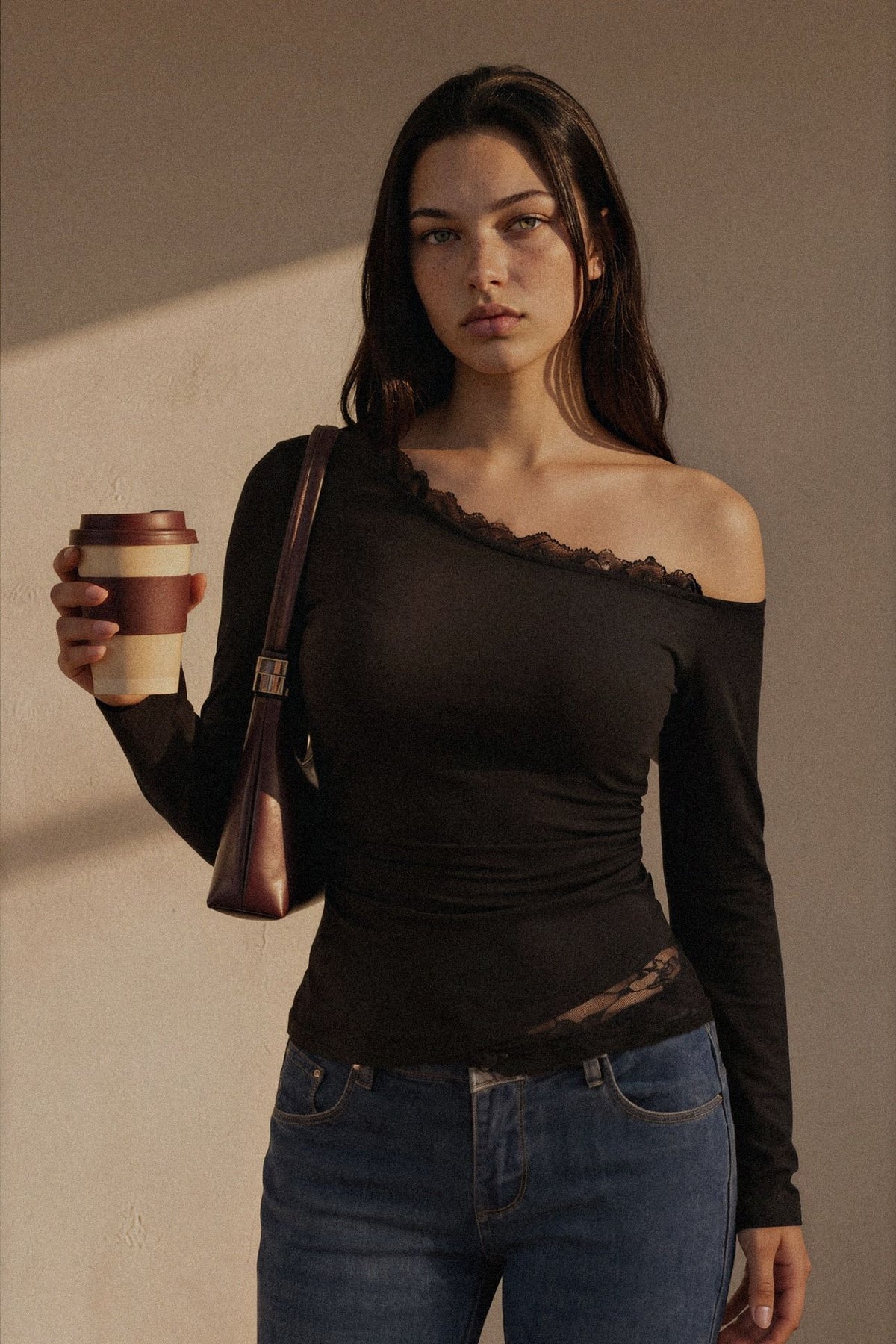 A woman wearing a black off-the-shoulder long-sleeve top with lace trim, paired with blue jeans, holding a brown and beige coffee cup and carrying a brown shoulder bag against a textured beige wall.