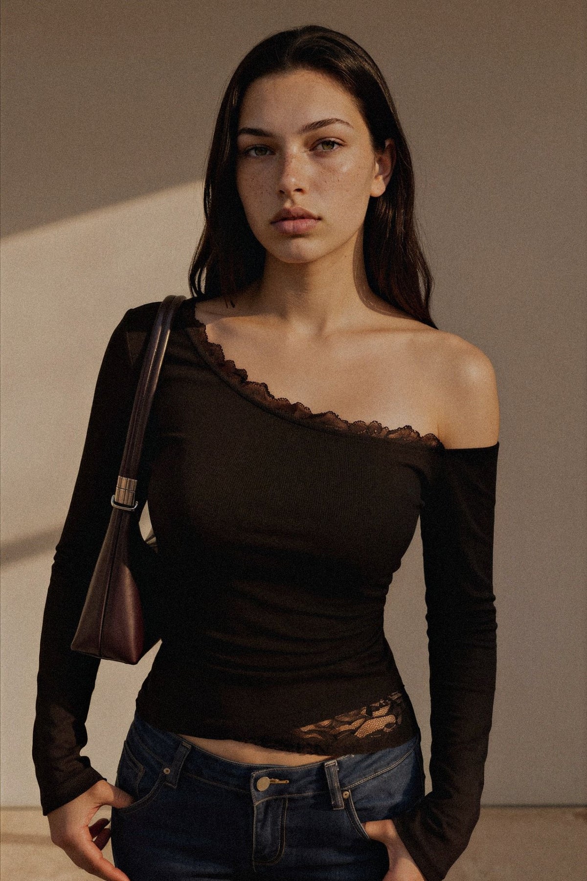 A woman wearing a dark brown off-the-shoulder long-sleeve top with lace trim, paired with blue jeans and carrying a brown shoulder bag, set against a neutral beige background.