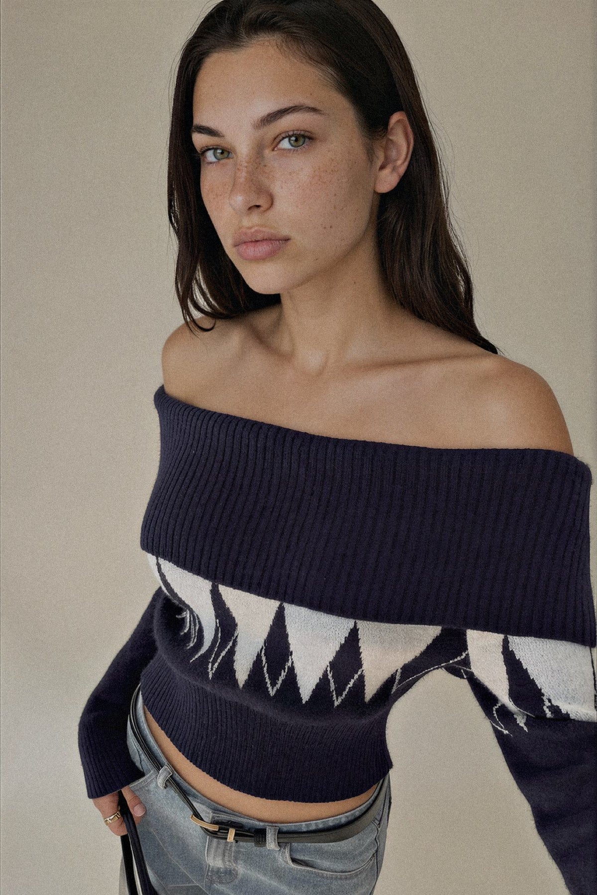 A woman wearing an off-the-shoulder navy blue knit sweater with a white geometric pattern across the chest and sleeves, paired with light-wash jeans, against a plain beige background.