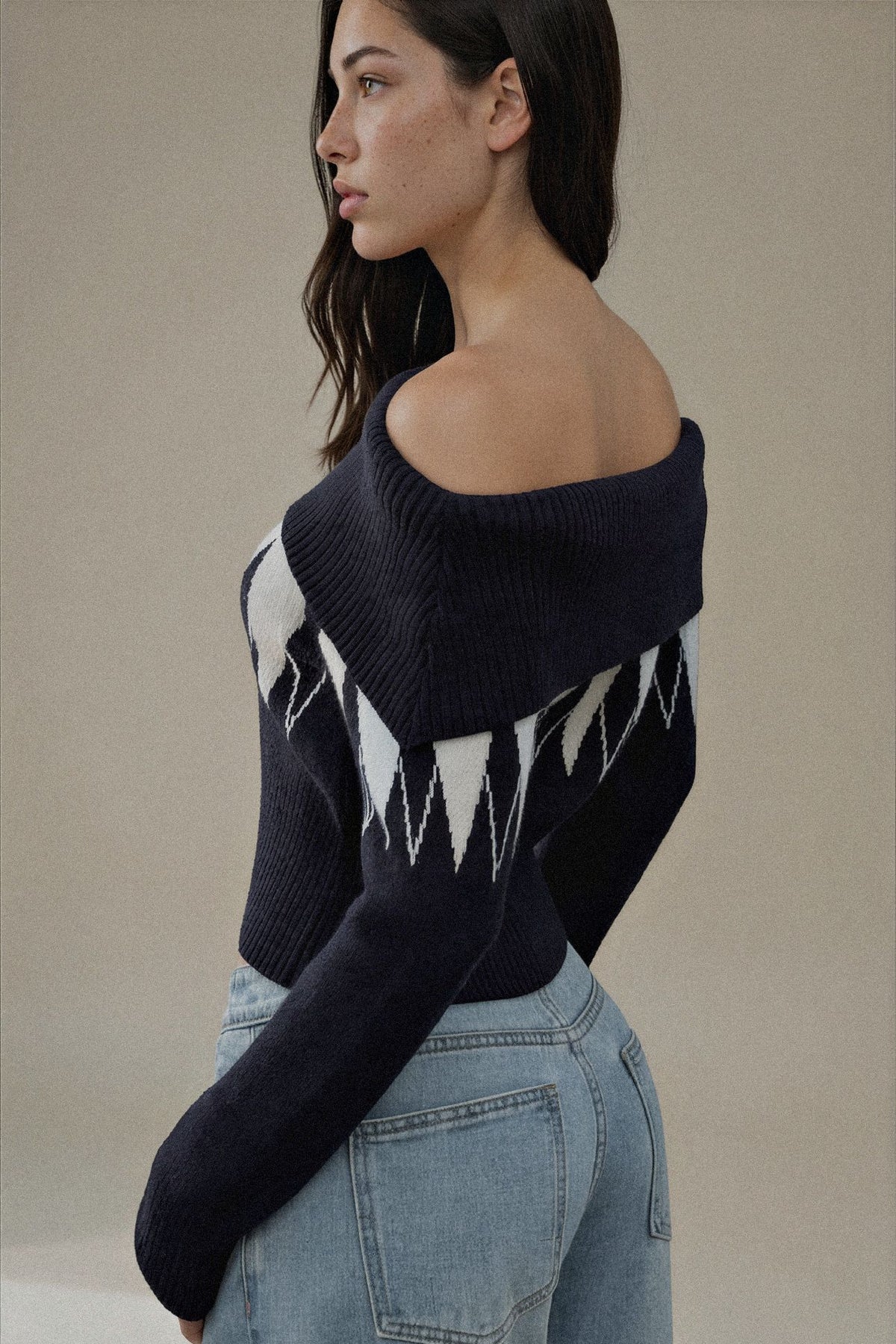 A woman wearing a dark navy off-the-shoulder knit sweater with a white geometric zigzag pattern on the front, paired with light blue jeans, photographed against a neutral beige background.