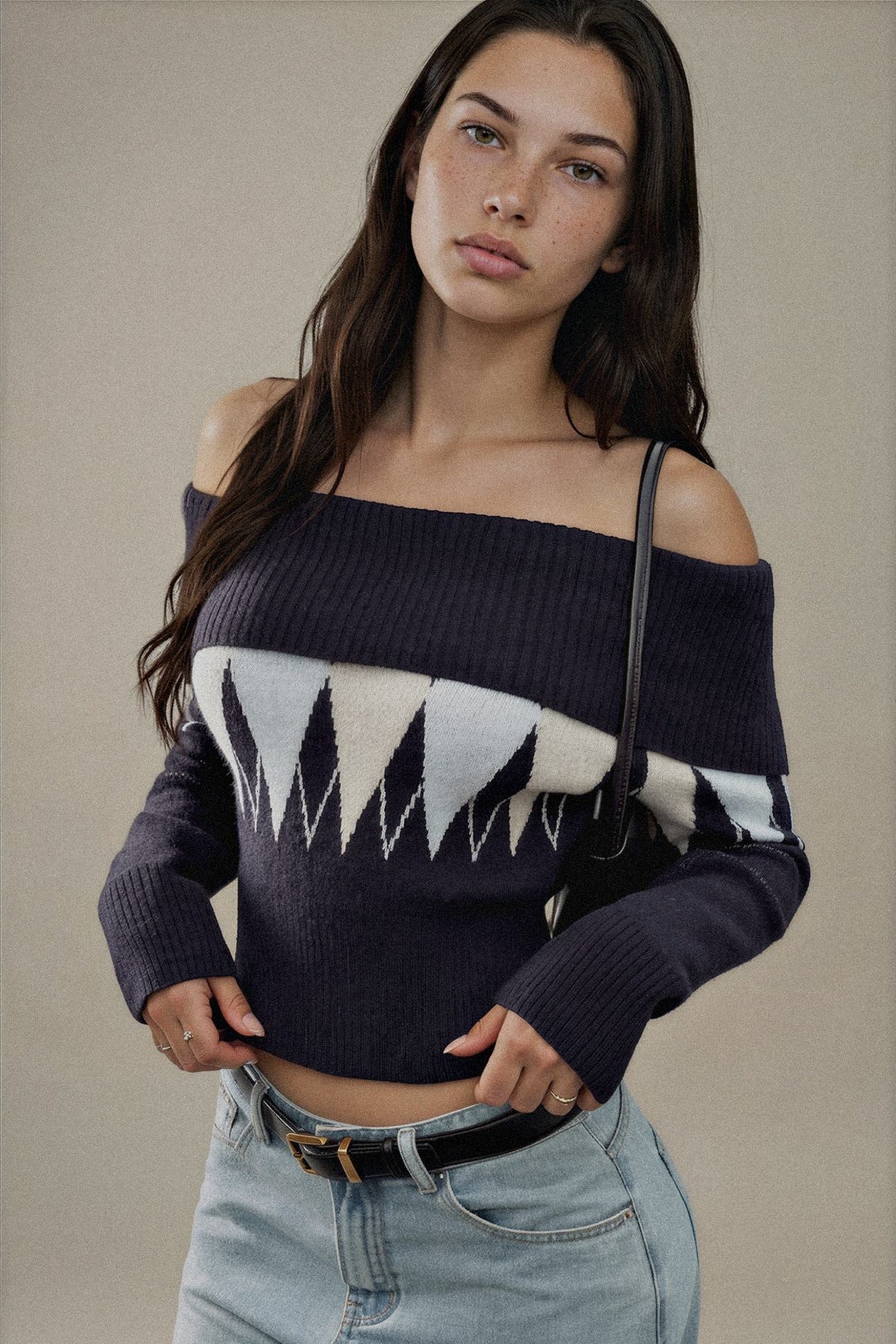 A woman wearing a dark navy off-the-shoulder knit sweater with a geometric white and light gray pattern across the chest, paired with light-wash jeans against a neutral beige background.