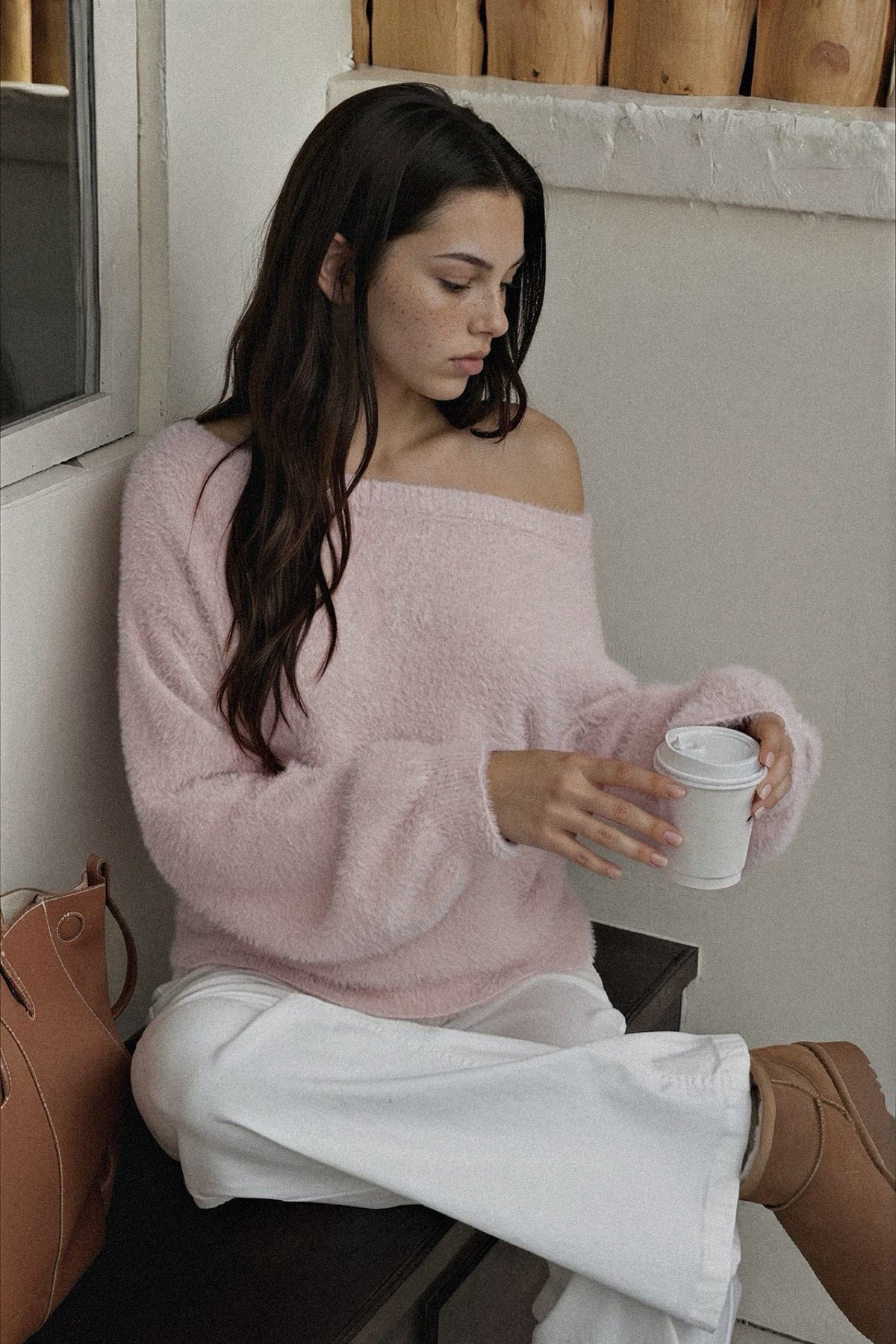 A woman sits on a bench wearing a fuzzy off-the-shoulder light pink sweater and white wide-leg pants, holding a white paper coffee cup in a neutral indoor setting.
