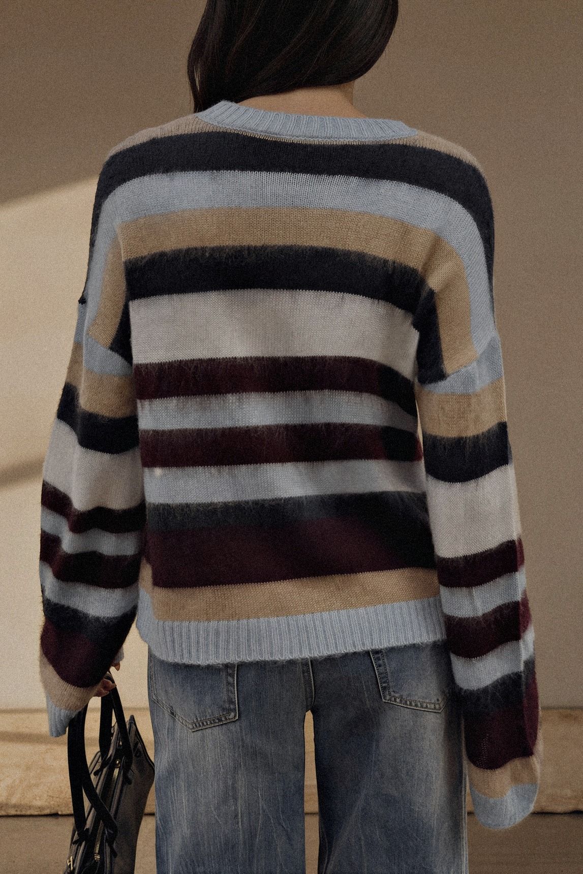 A person shown from the back wearing a fuzzy striped knit sweater with horizontal bands of light blue, beige, dark navy, burgundy, and cream, paired with blue jeans.
