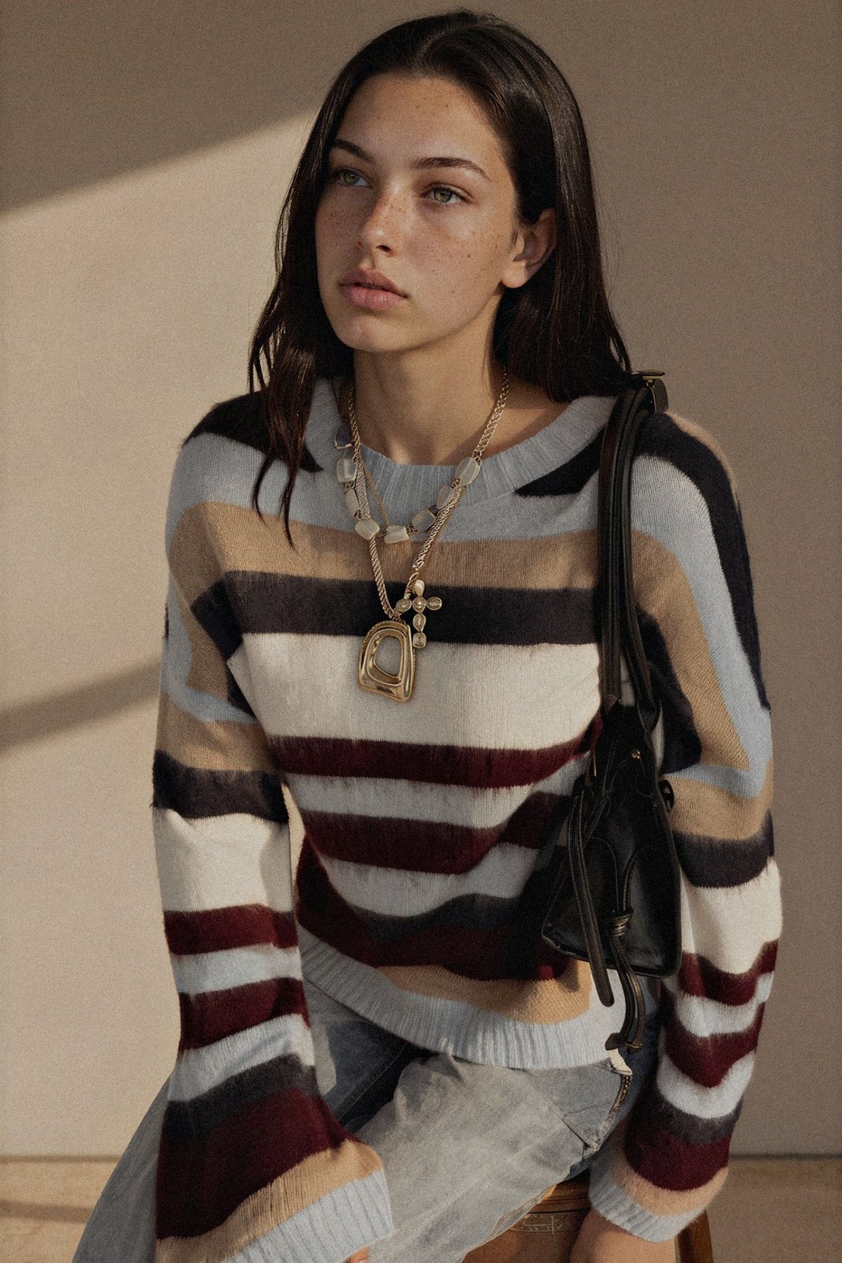 A woman wearing a fuzzy knit sweater with horizontal stripes in cream, beige, brown, navy, and burgundy, paired with light-wash jeans and layered gold necklaces, holding a black shoulder bag.
