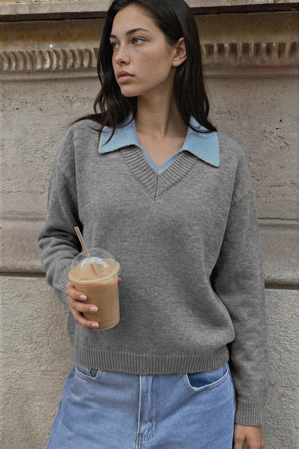 A woman wearing a gray knit sweater with a light blue collar detail, holding an iced coffee in front of a textured stone wall.