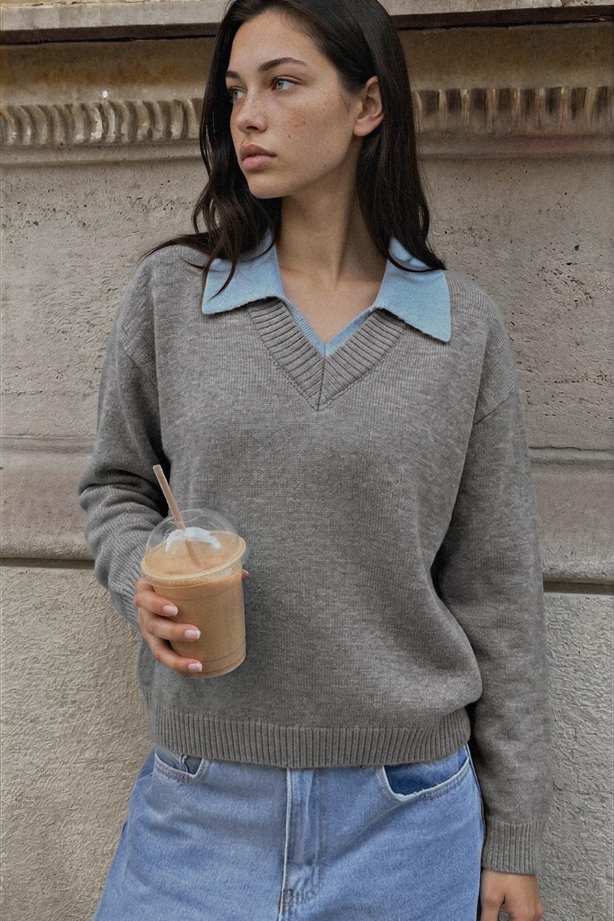 A woman wearing a gray knit sweater with a light blue collar detail, holding an iced coffee in front of a textured stone wall.