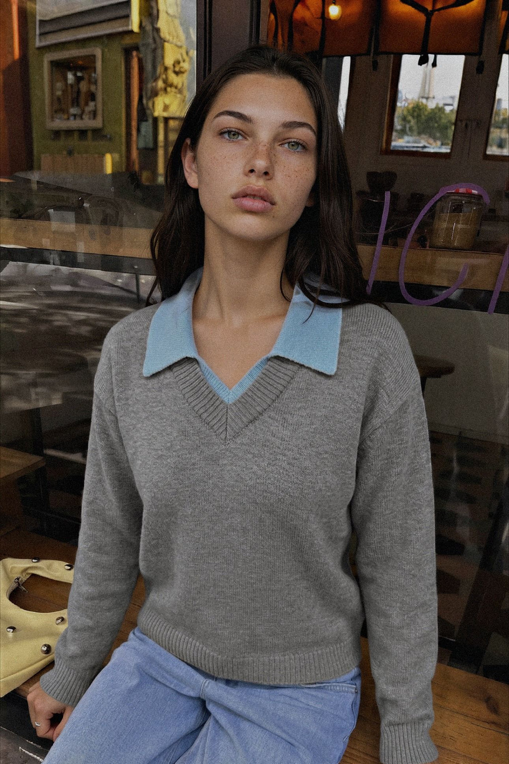 A woman wearing a gray knit sweater with a light blue collar, paired with light blue jeans, seated in a warm, rustic indoor setting.