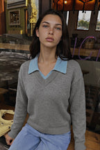 A woman wearing a gray knit sweater with a light blue collar, paired with light blue jeans, seated in a warm, rustic indoor setting.