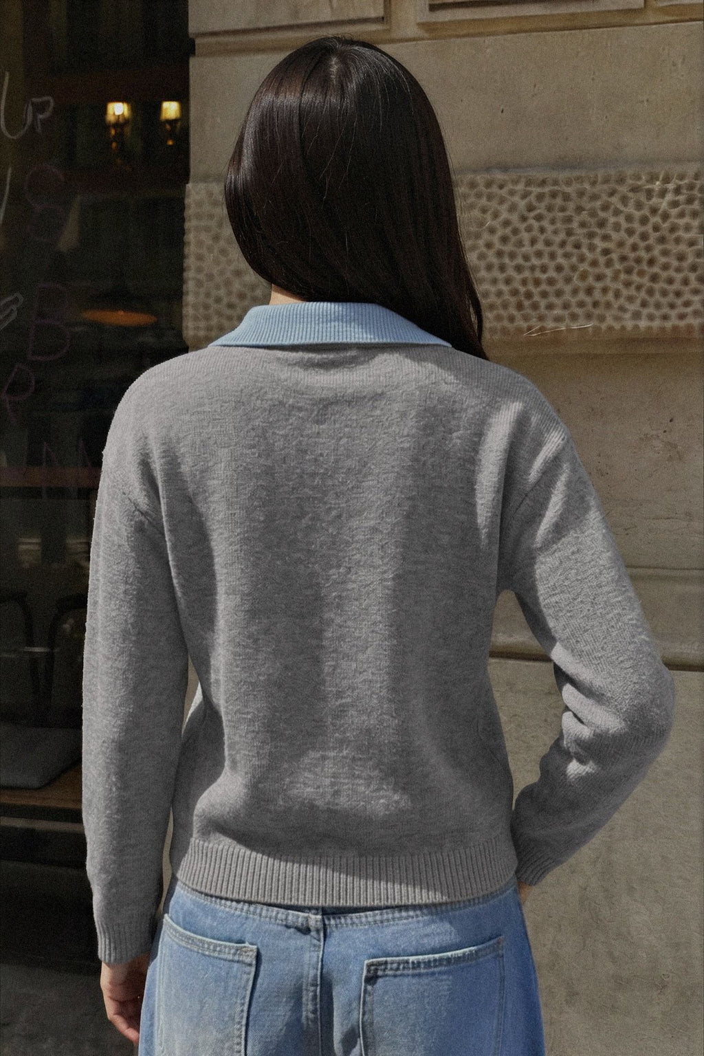 A woman viewed from behind wearing a light gray knit sweater with a light blue collar, paired with light blue jeans, standing outdoors against a stone wall.