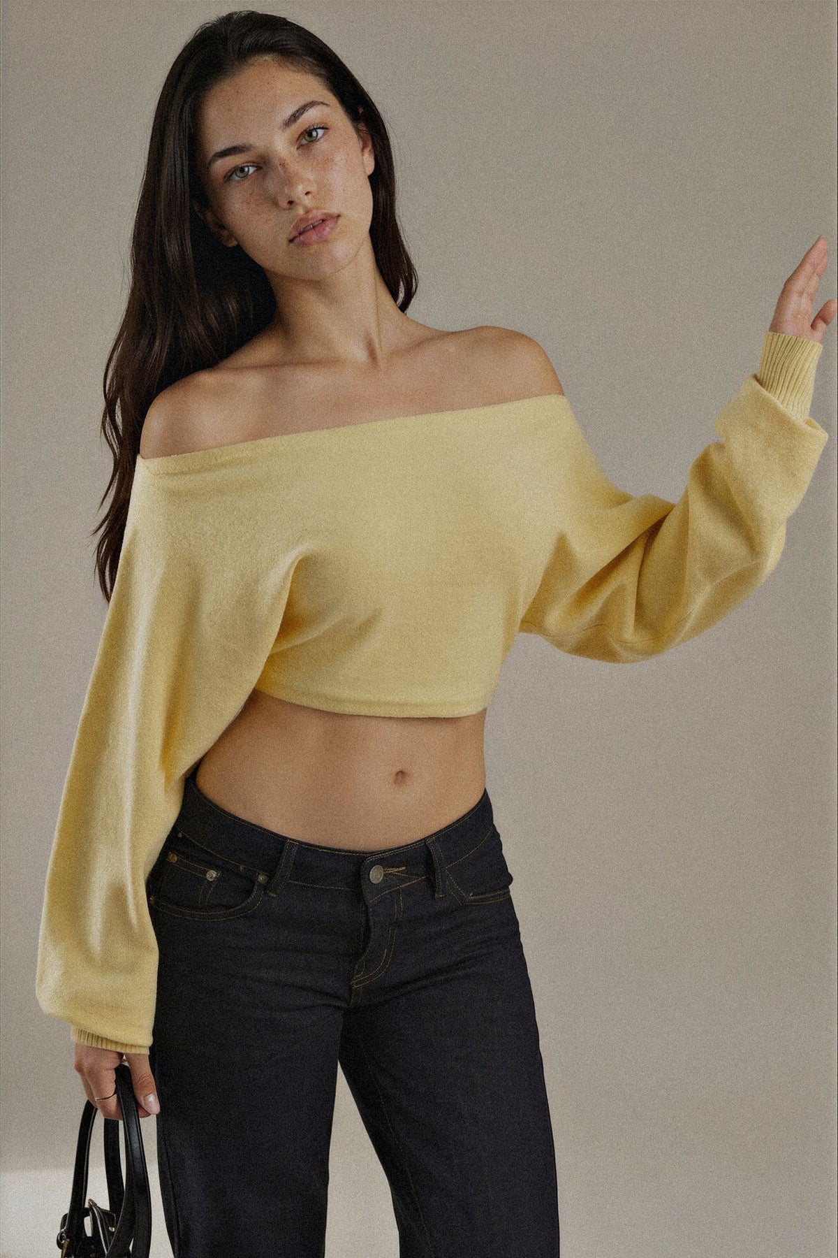 A woman modeling a pale yellow off-the-shoulder knit sweater with long balloon sleeves, paired with dark blue jeans, against a plain beige studio background.