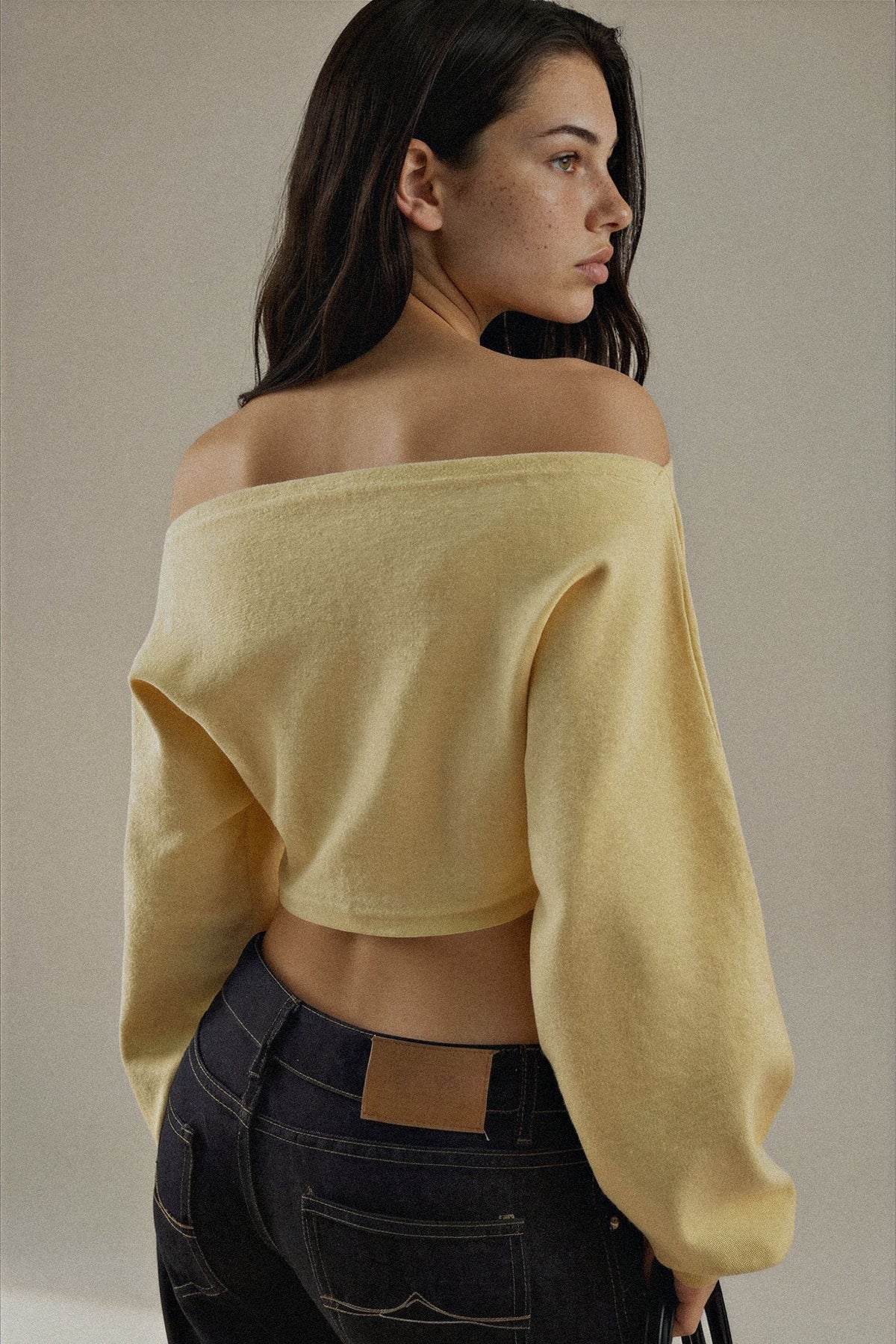 A woman wearing a pale yellow off-the-shoulder knit crop top and dark blue jeans, photographed against a plain warm beige studio background.