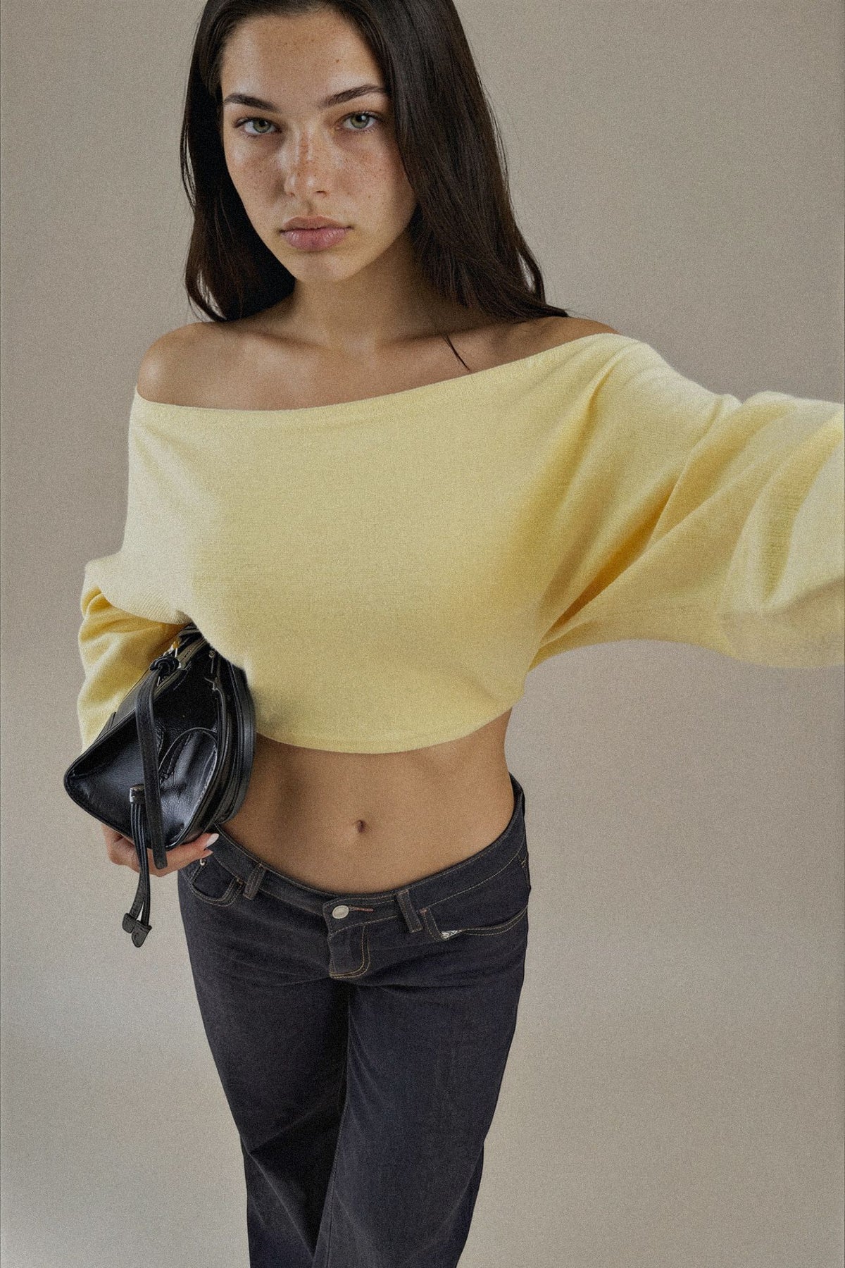 A woman modeling a pale yellow off-the-shoulder knit crop top with wide draped sleeves, paired with dark jeans and holding a black crossbody bag against a plain beige studio background.