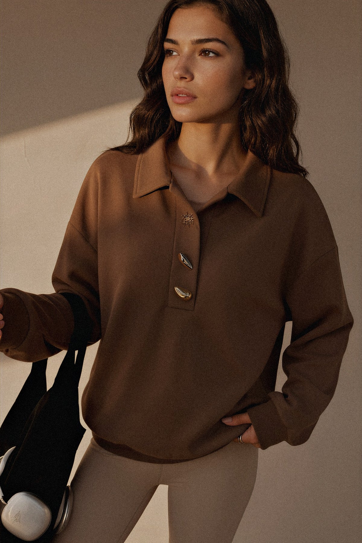 A woman wearing a brown long-sleeve knit polo-style top with decorative buttons, paired with light beige leggings, holding a black tote bag against a warm beige background.
