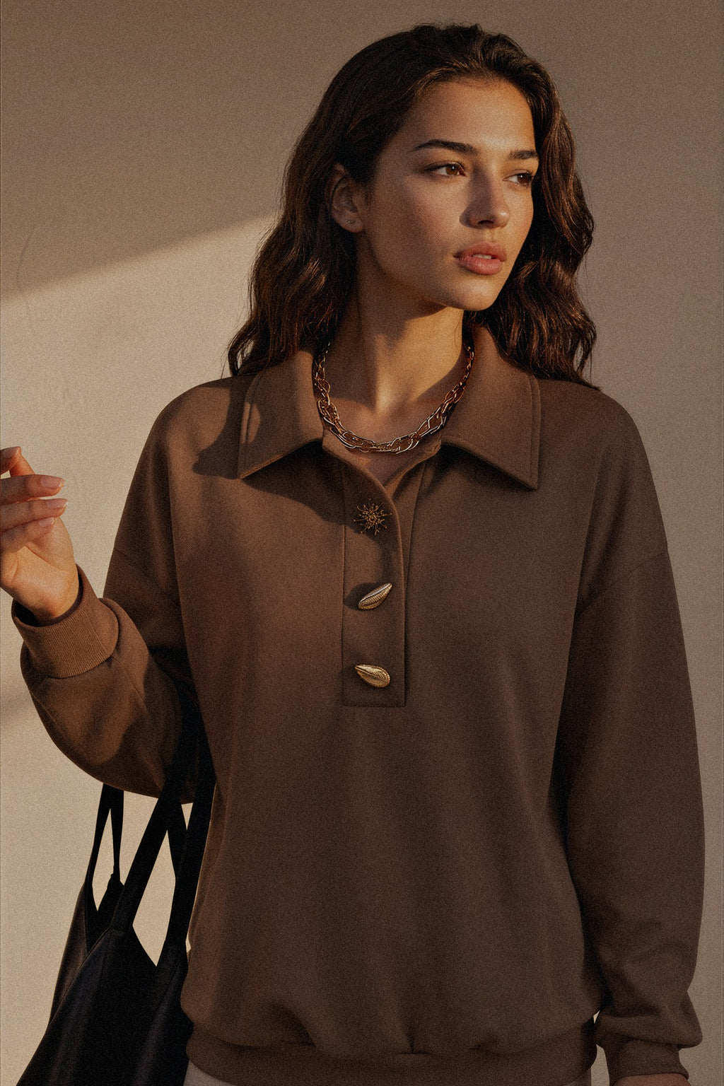 A woman wearing a brown long-sleeve collared knit top with three decorative shell-like buttons on the front, styled with layered chain necklaces and a black tote bag, set against a warm beige wall with dramatic side lighting.