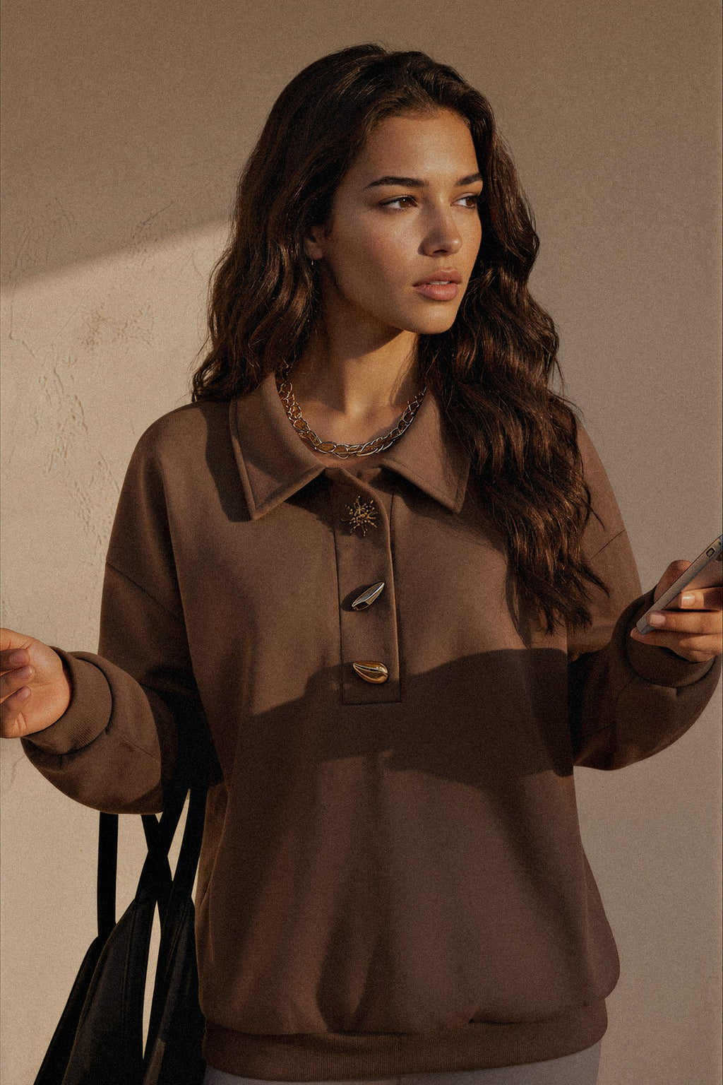 A woman wearing a brown long-sleeve polo sweatshirt with a buttoned placket and decorative chest details, standing against a textured beige wall.